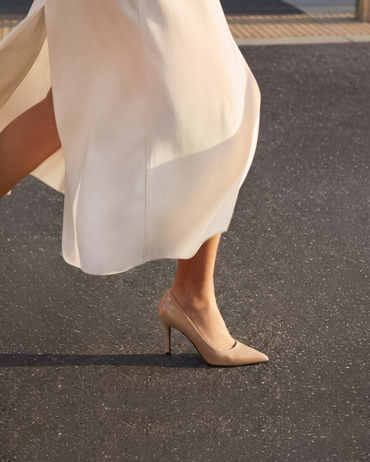 Beige high heeled shoes worn with a flowing white dress on a dark surface.