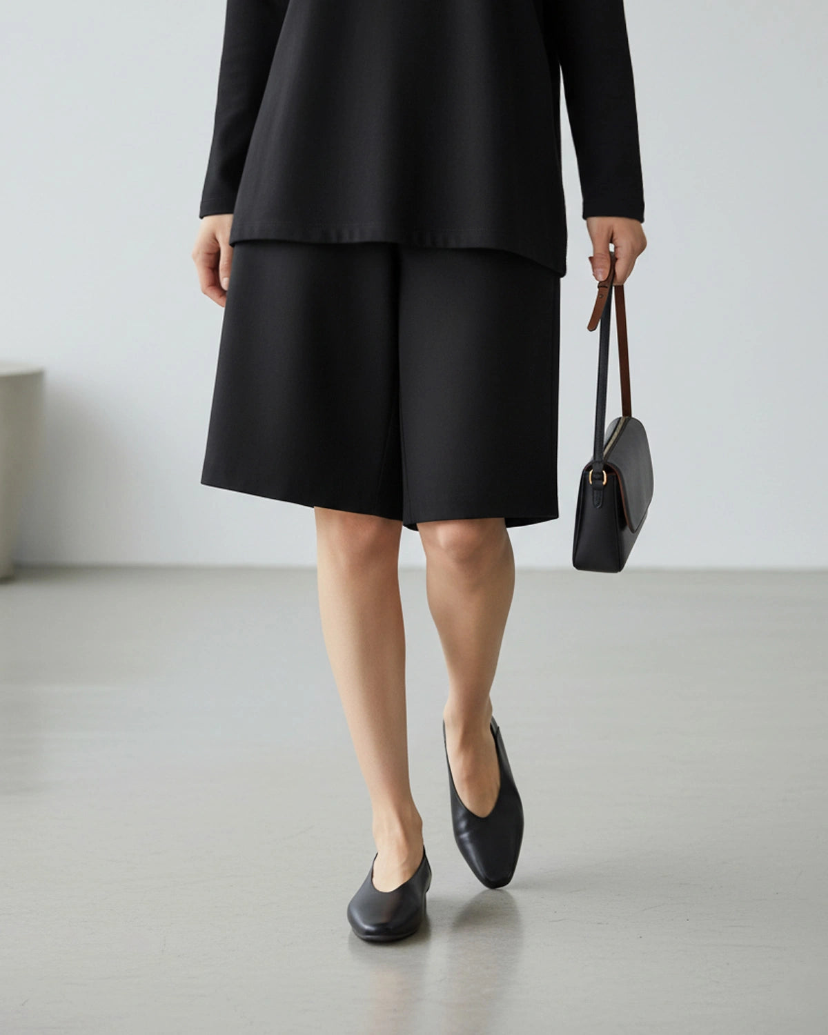 Woman wearing a black outfit with black flat shoes, holding a handbag. 