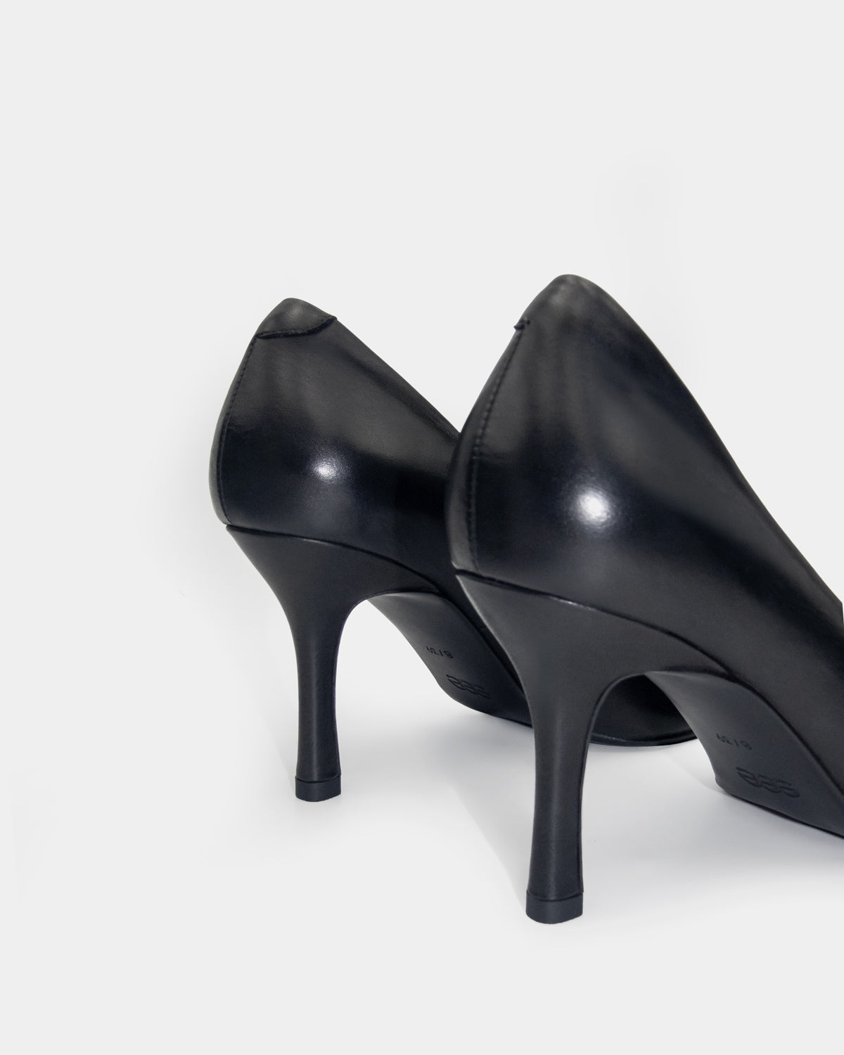 A pair of black leather high-heeled pumps shown from a rear three-quarter angle on a light gray studio background.