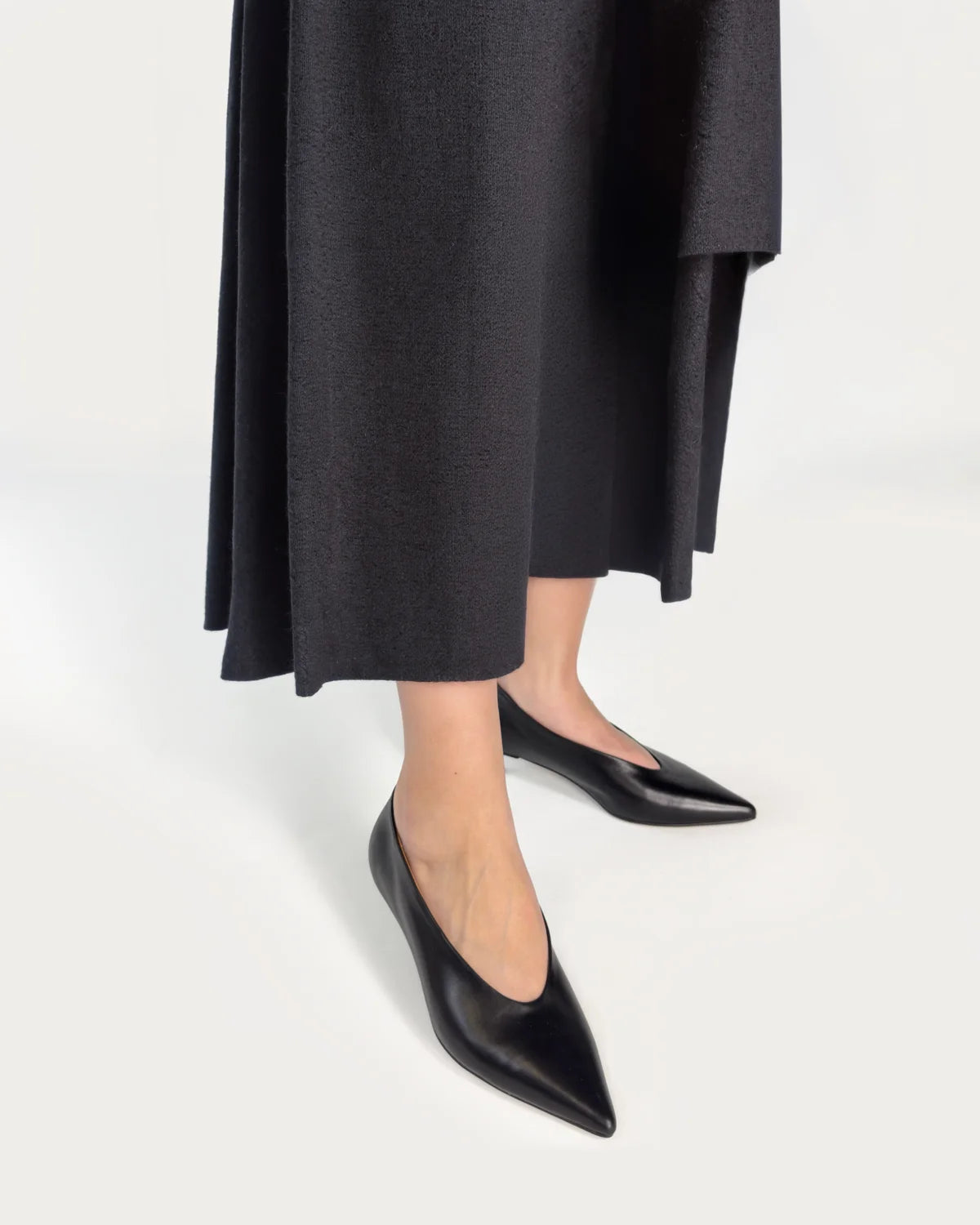 Person wearing black pointed-toe kitten heels and a long black coat on a white background