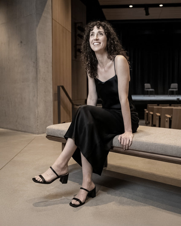 Woman in a black dress and black sandals sitting on a bench in an indoor setting