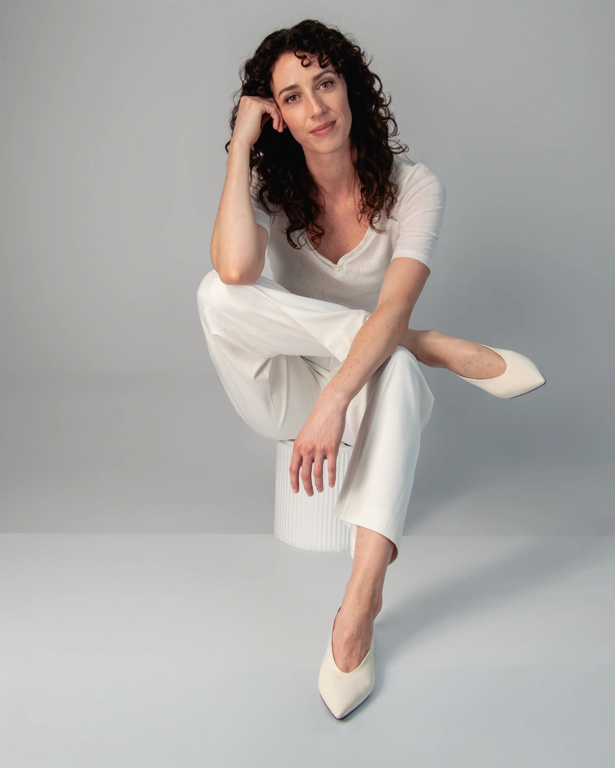 Woman in a white outfit and white heels sitting on a white stool against a grey background