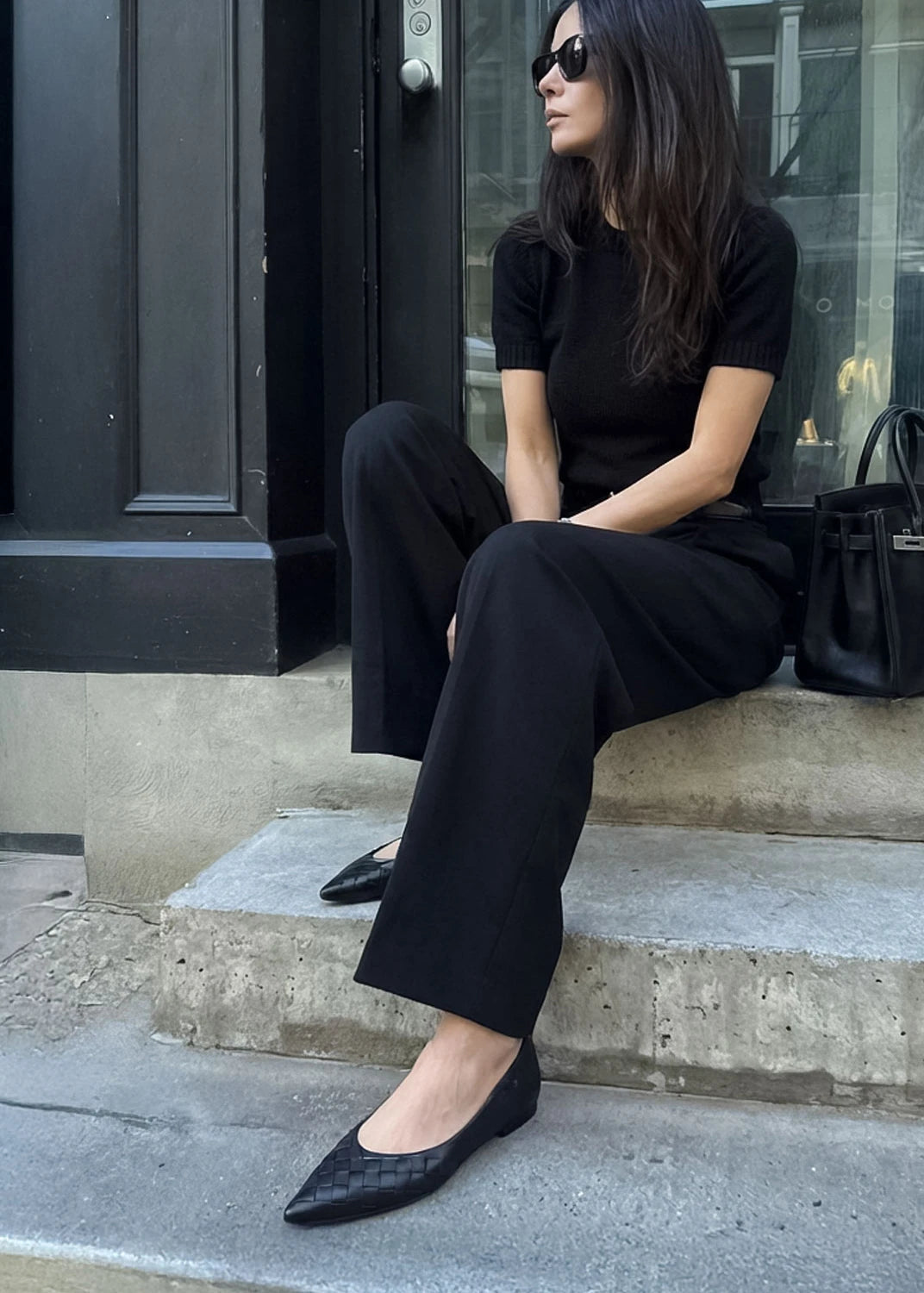 Woman in black outfit sitting on steps wearing black flat shoes.