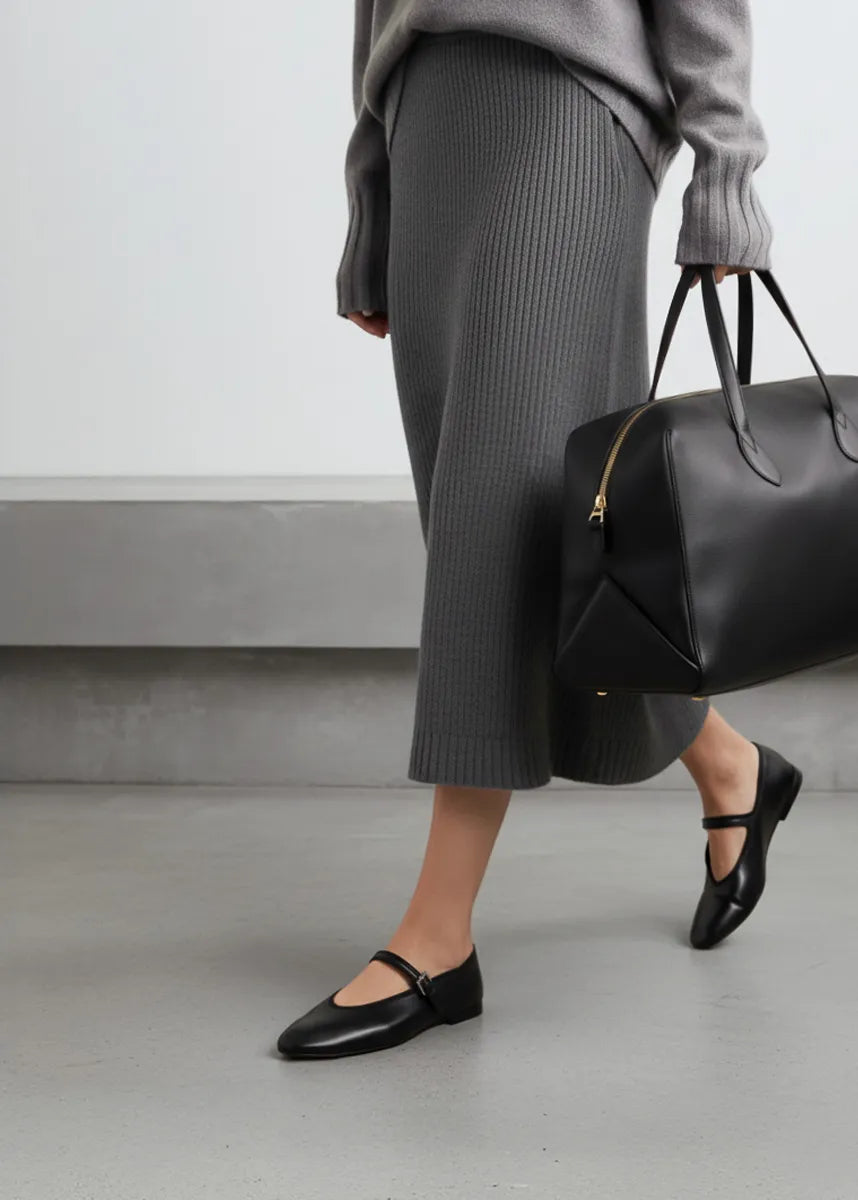 woman holding a black handbag wearing a grey outfit, and black ballet flat shoes.