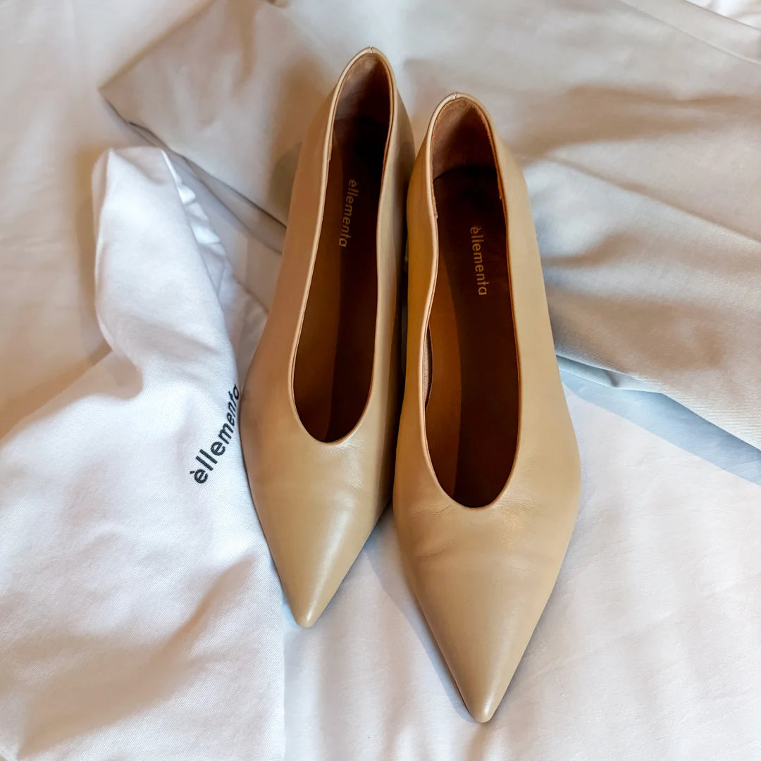 A pair of beige pointed-toe pumps with a smooth leather finish, displayed on a white fabric surface with a white branded dust bag.
