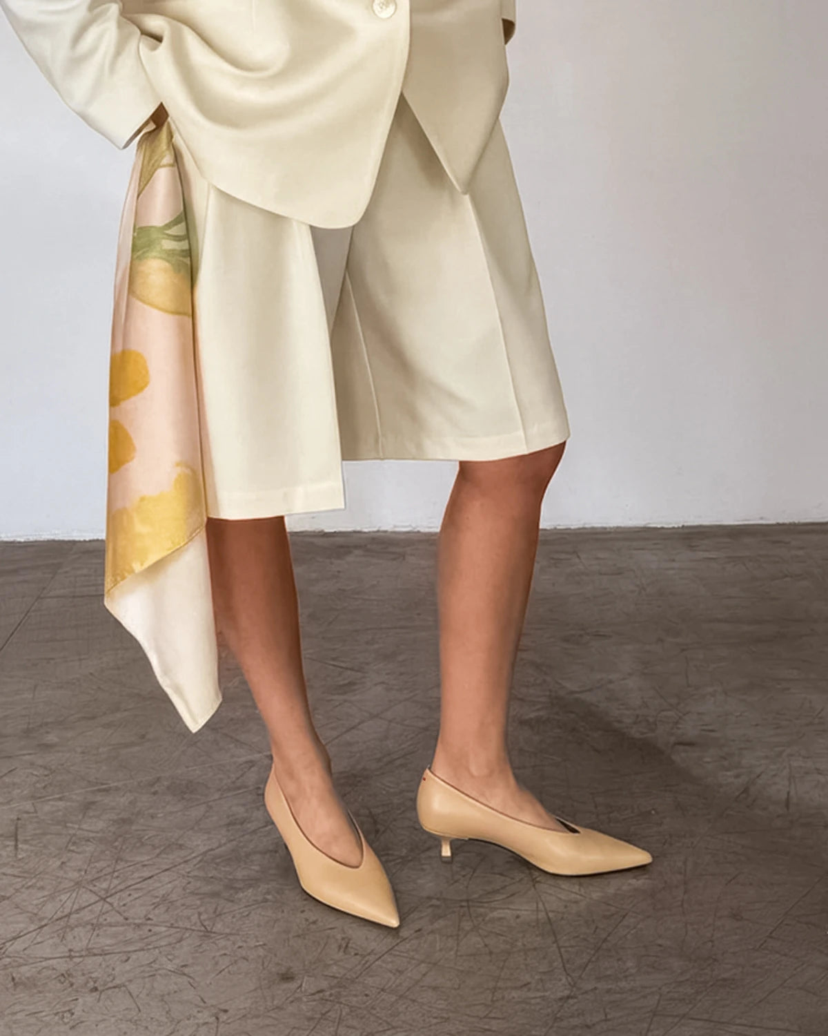 Woman wearing a beige pointed toe shoes and outfit with a floral scarf, standing on a textured floor.