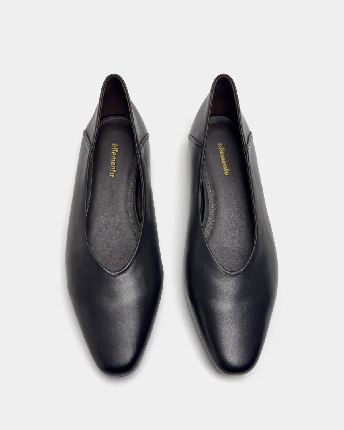 A pair of black leather slip-on flats with round toes and a low block heel, shown from a top-down view on a light gray background. 