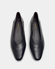 A pair of black leather slip-on flats with round toes and a low block heel, shown from a top-down view on a light gray background. 