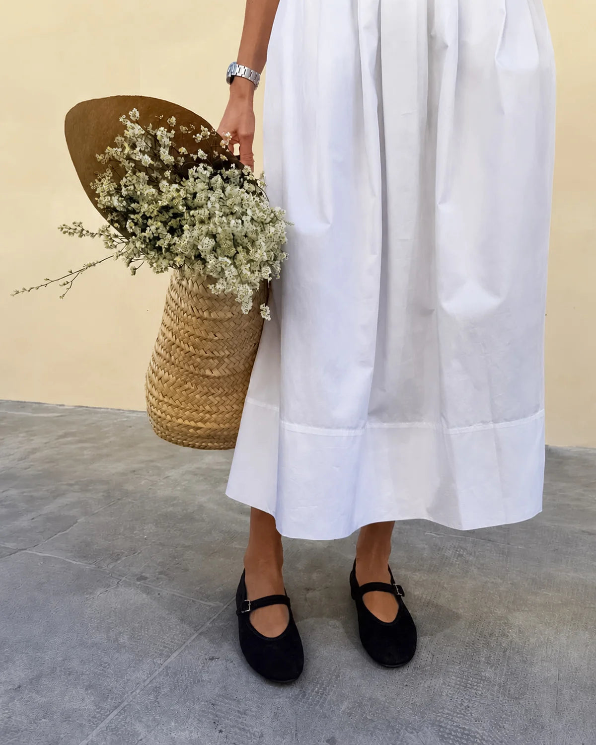A person wearing a long white midi skirt with a pleated waist and double hem, styled with black Mary Jane flats and holding flowers 