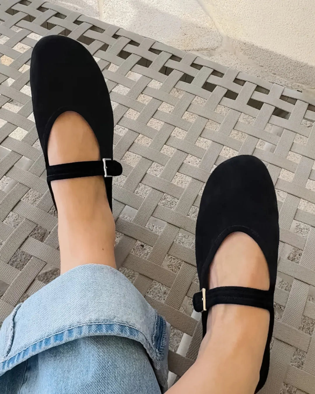 A person wearing a pair of black flat Mary Jane shoes with ankle straps and small silver buckle details, paired with light blue jeans, sitting on a woven patio chair.
