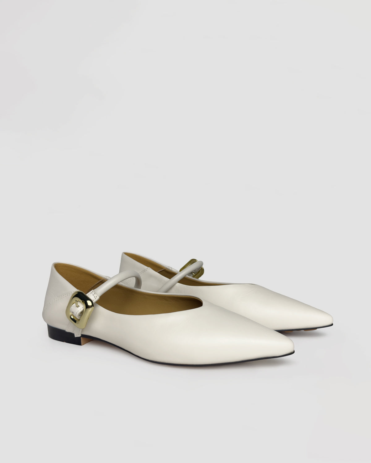 A pair of off-white leather Mary Jane flats with pointed toes and gold-tone buckle straps, displayed on a neutral light gray background.