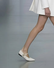 A close-up of a model's legs walking on a runway, wearing a pair of pointed-toe Mary Jane flats in off-white with a small gold buckle and a black heel.