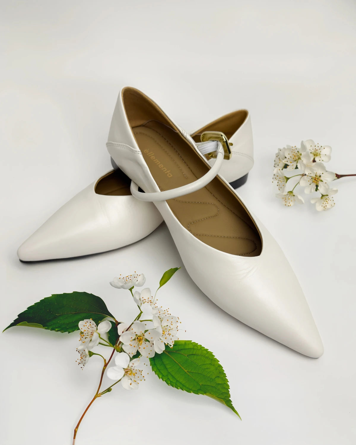A pair of white pointed-toe Mary Jane flats with a single strap and a small gold buckle, styled on a white background with green leaves and white blossoms.