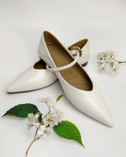 A pair of white pointed-toe Mary Jane flats with a single strap and a small gold buckle, styled on a white background with green leaves and white blossoms.