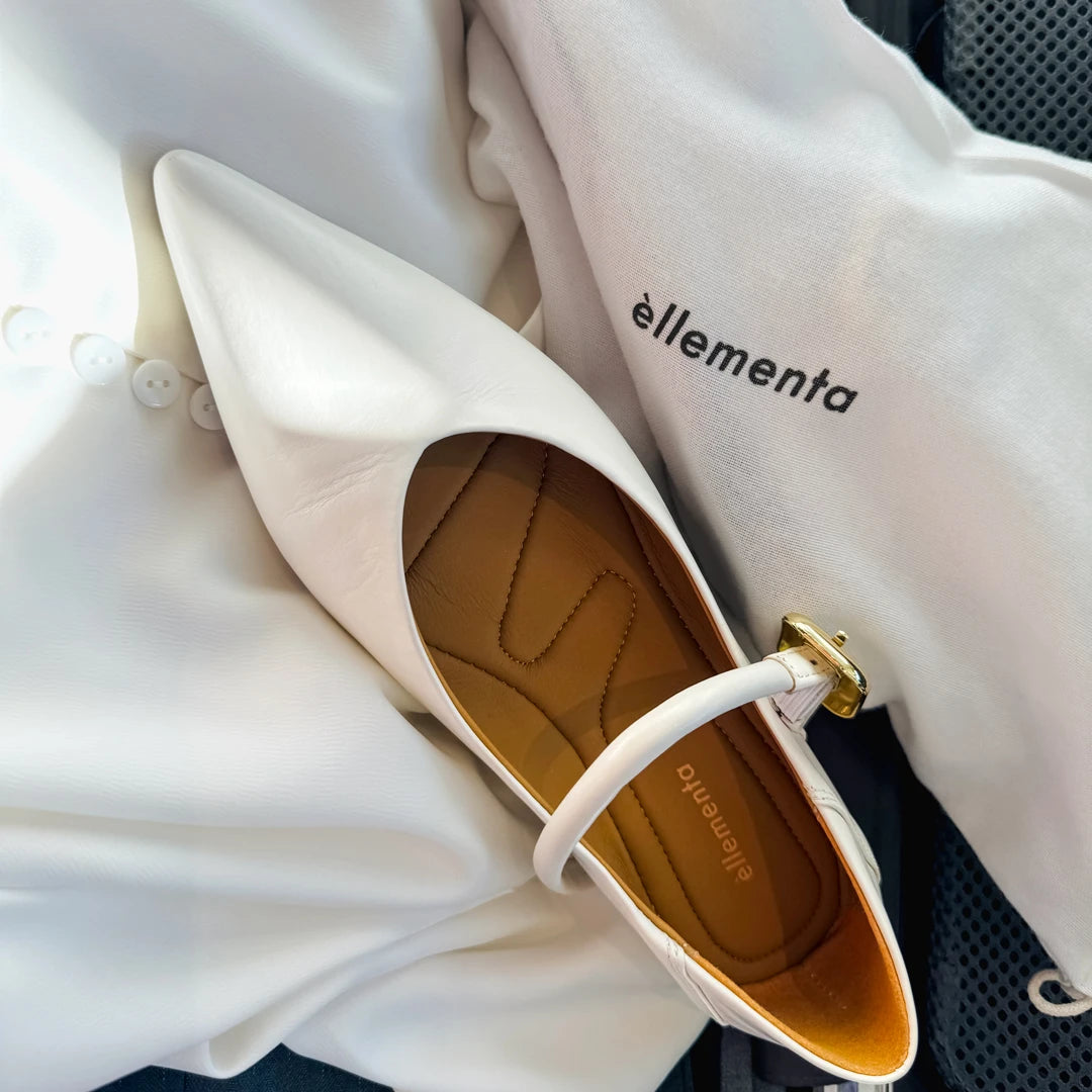 A white pointed-toe flat shoe with a small gold-tone buckle and white strap, shown resting on white fabric next to a white dust bag printed with the brand name 'ellementa'.