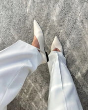 White pointed-toe flat shoes worn with white wide-leg trousers, shown from a top-down view on a gray stone pavement.