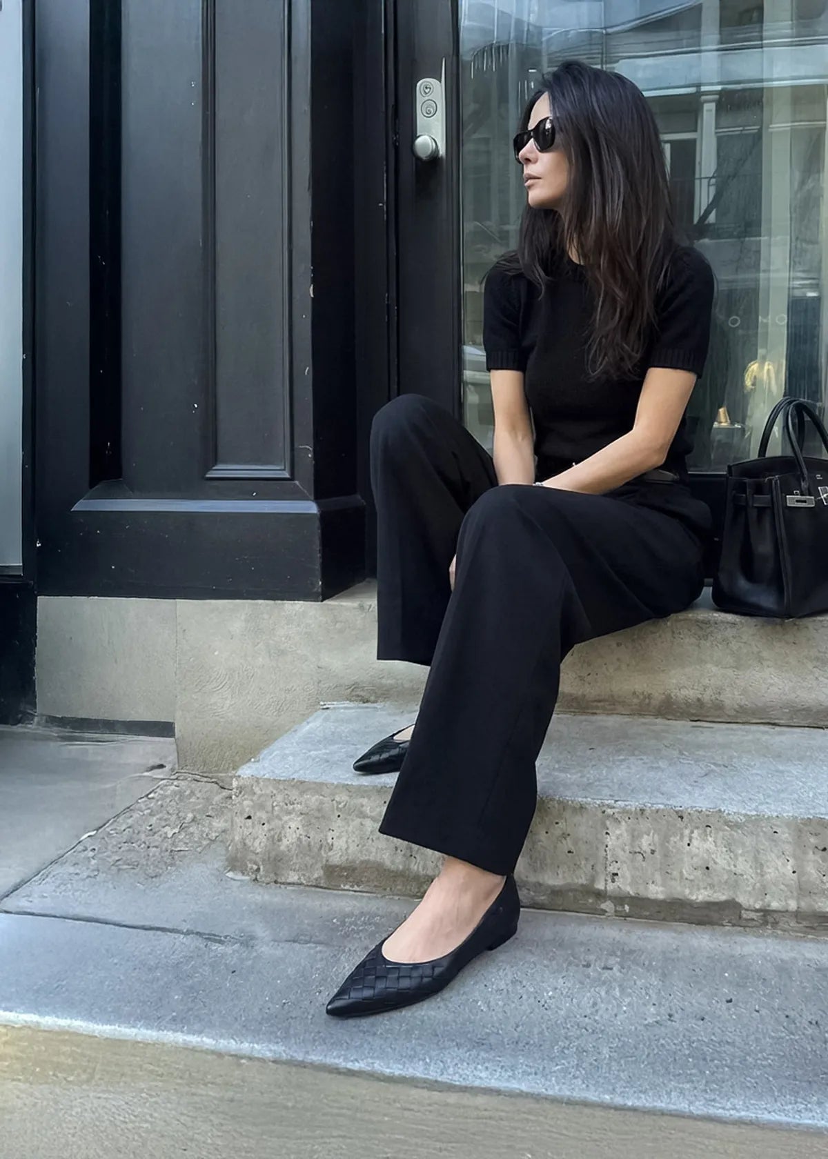 Woman in black outfit sitting on steps black leather flats and holding a black handbag.