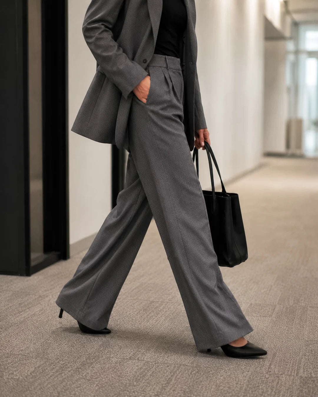 A person walking in a hallway wearing a tailored gray blazer and  wide-leg trousers and black heeled pointed-toe shoes
